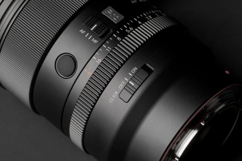 A close-up view of a camera lens barrel featuring an aperture ring with f-stop markings, an AF/MF switch, a customizable button, and a click on/off switch.