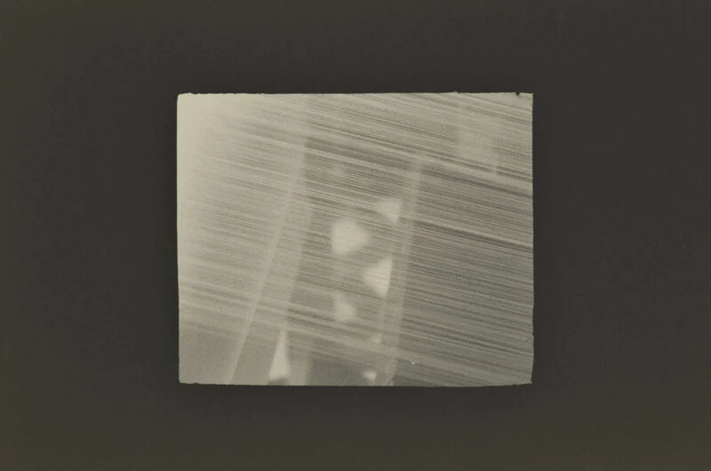 A rectangular, sepia-toned print with a textured, streaked appearance features faint, geometric shapes layered behind diagonal lines.