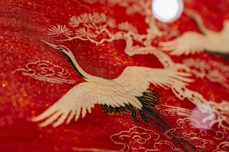 A close-up of red fabric featuring gold-threaded embroidery of cranes, pine branches, and stylized clouds.