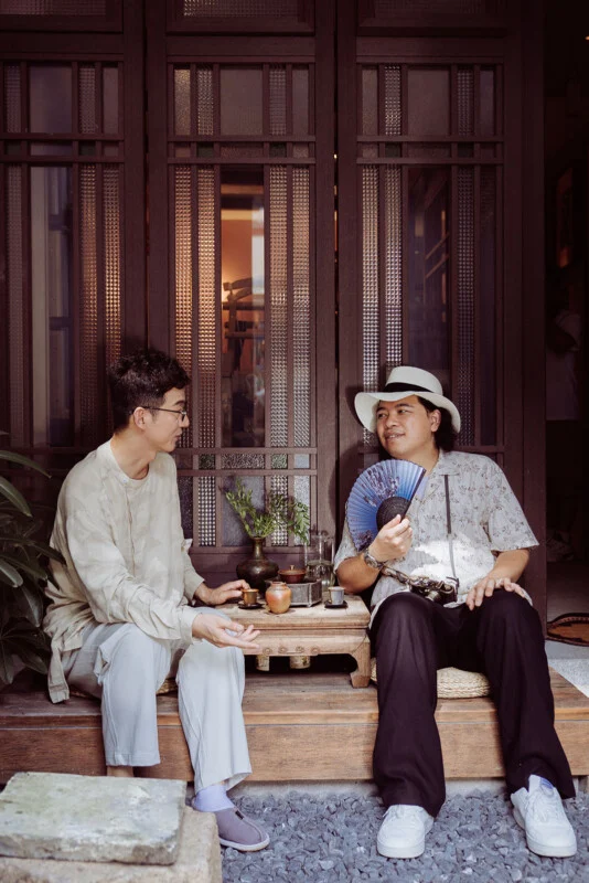 Two men sit on a wooden porch in front of a dark wooden door, talking while sharing tea at a small table.