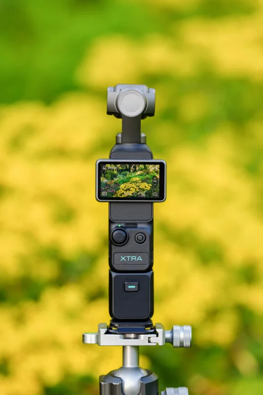 A handheld gimbal camera with a digital screen displaying a garden scene is mounted on a tripod against a blurred yellow and green background.