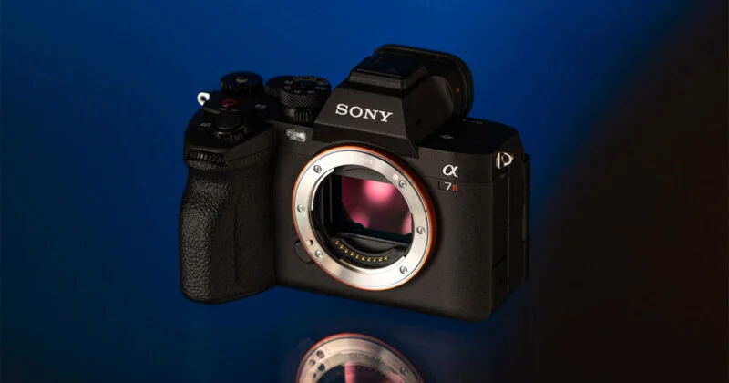 A black Sony Alpha 7R mirrorless camera body sits on a reflective surface against a blue background.