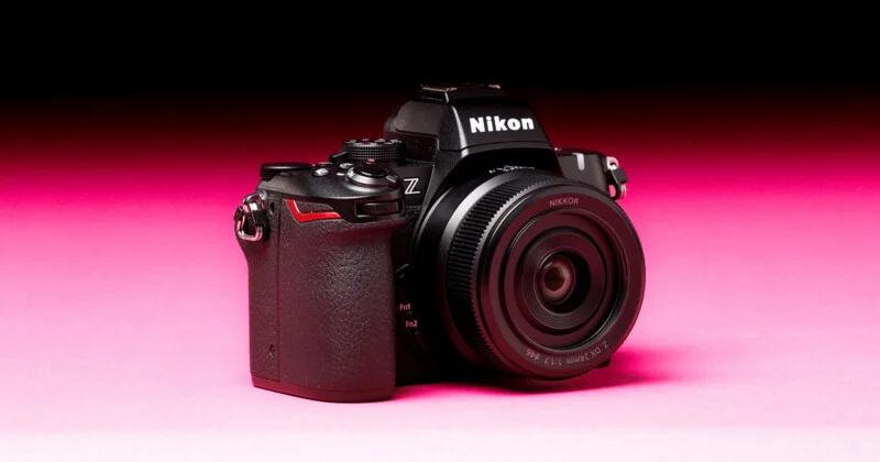 A black Nikon Z mirrorless camera with a 24mm lens sits on a pink surface against a dark background.