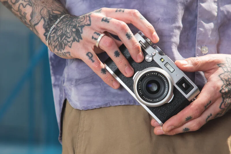 A person with tattooed hands holds a silver and black Fujifilm camera against a light purple shirt and tan pants.
