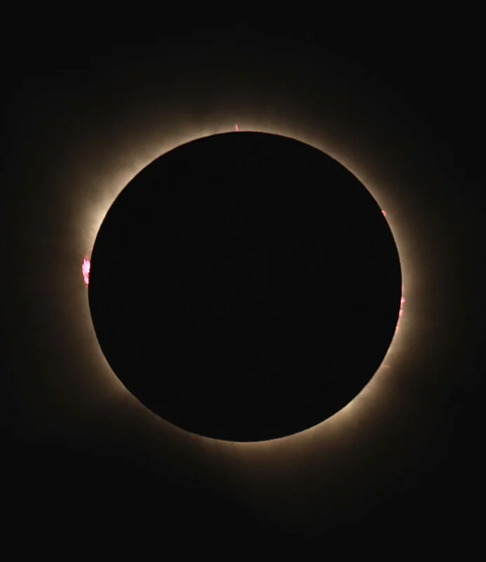 A total solar eclipse shows the moon silhouetted against the sun, with a glowing corona and bright red solar prominences.