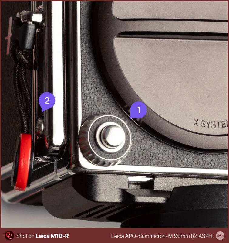 A close-up view of a camera body showing a lens release button labeled 1 and a strap lug labeled 2, with text at the bottom indicating it was shot on a Leica M10-R.