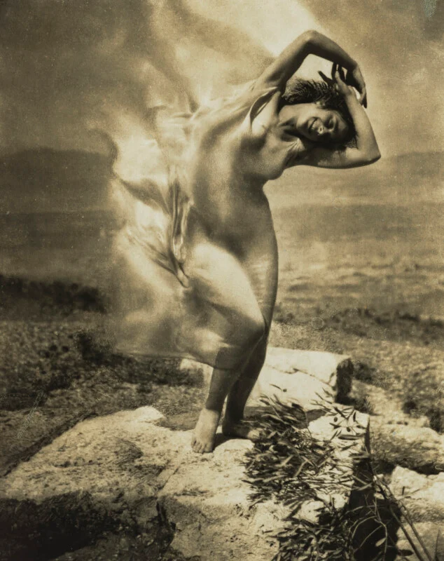 A woman with arms raised above her head dances on a stone platform, her sheer garment blurred by motion against a landscape.