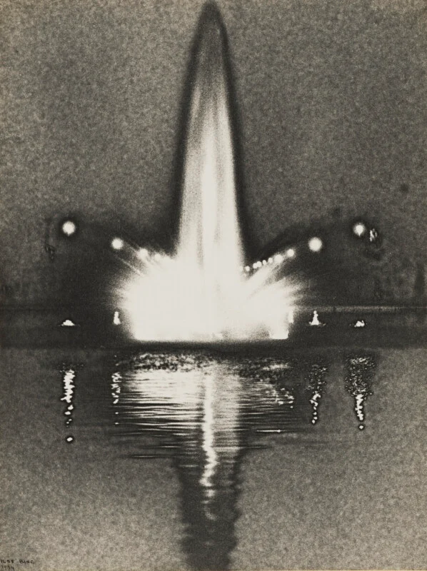 A black-and-white photograph shows a brightly illuminated fountain shooting a tall jet of water into the night air, with its reflection shimmering on the dark water below.