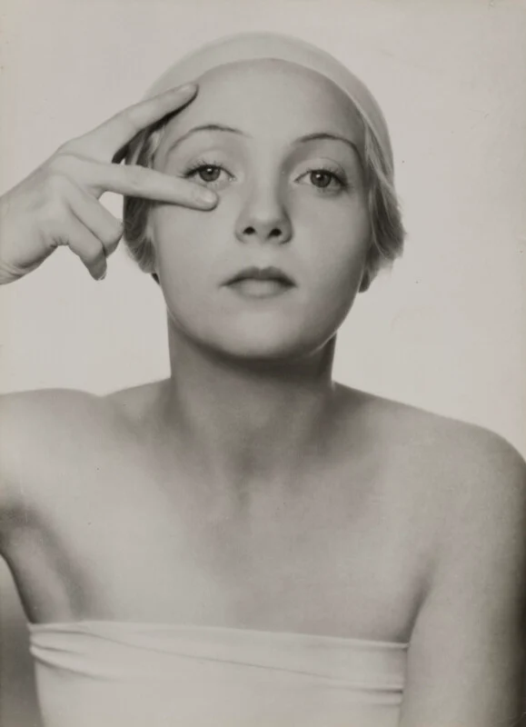 A black-and-white portrait of a woman wearing a swim cap, holding two fingers to her right eye.