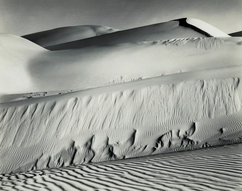 A black-and-white landscape of rolling sand dunes with prominent wind-swept ripples and deep shadows.