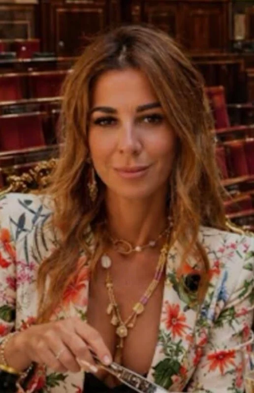 A woman with long, wavy brown hair wears a floral-patterned blazer and layered gold necklaces while holding a piece of cutlery.