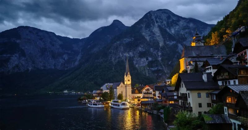 A lakeside village with a tall church spire and illuminated buildings sits at the base of steep, dark mountains at dusk.