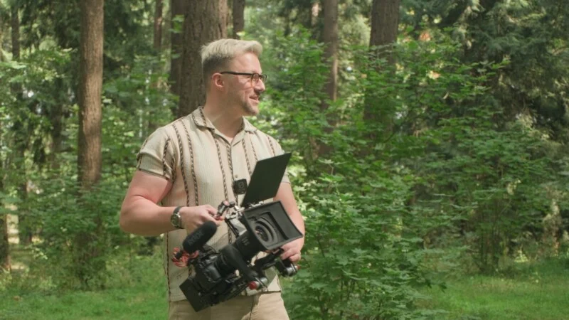 A man with blonde hair and glasses stands in a forest, holding a professional video camera with a microphone and matte box attached.