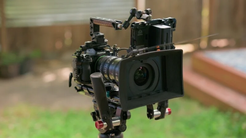 A professional cinema camera rig mounted on a tripod, featuring a lens hood, a top handle, and an external monitor attached to the back.