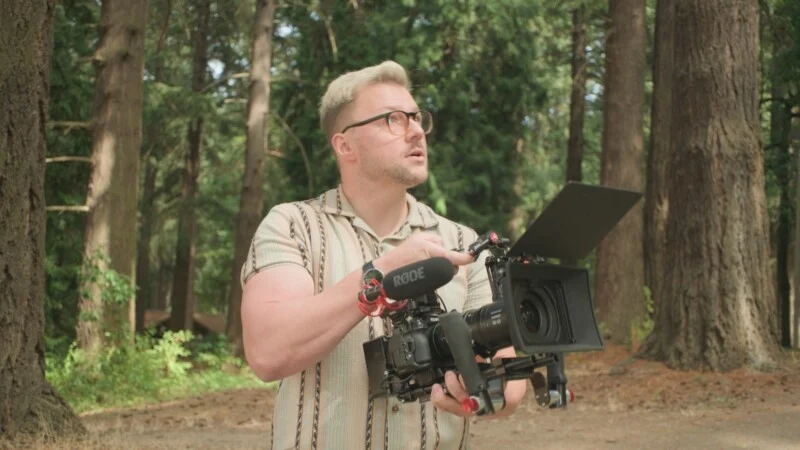 A man with blonde hair and glasses holds a professional video camera with a microphone attached while standing in a forest.