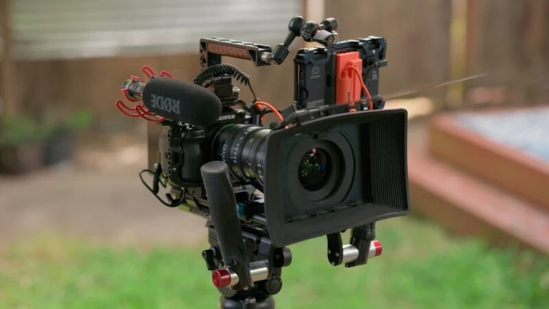 A professional cinema camera rig mounted on a tripod, featuring a lens hood, a Rode microphone, and an Atomos external monitor.