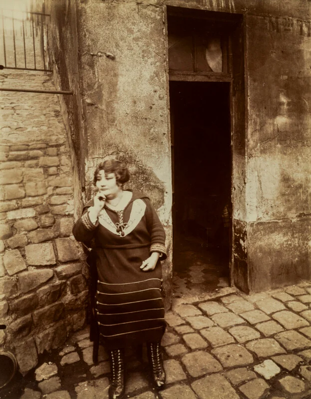 A woman with short, dark hair sits on a chair in a cobblestone alleyway, resting her chin on her hand while wearing a dark dress with a white collar and lace-up boots.