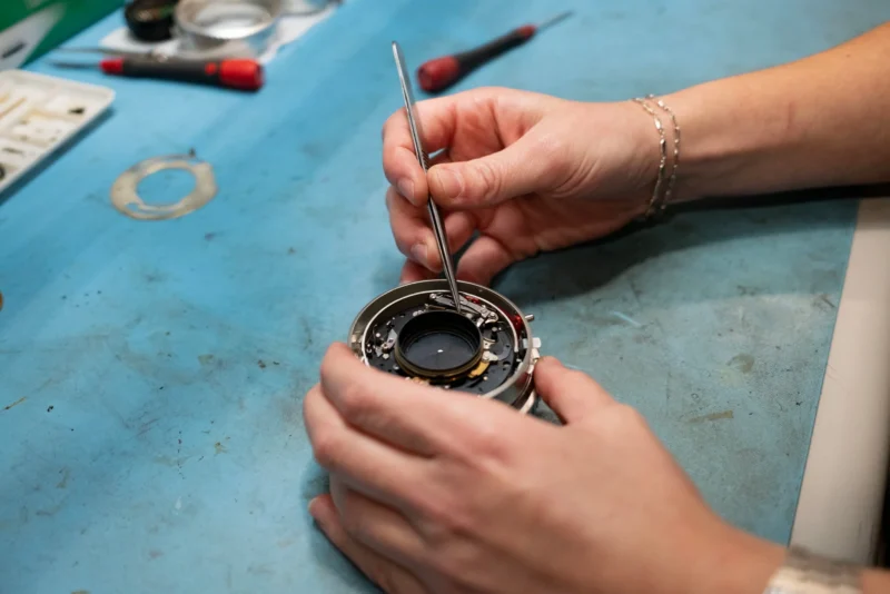 A person uses tweezers to adjust the internal mechanism of a disassembled camera lens on a blue workbench.