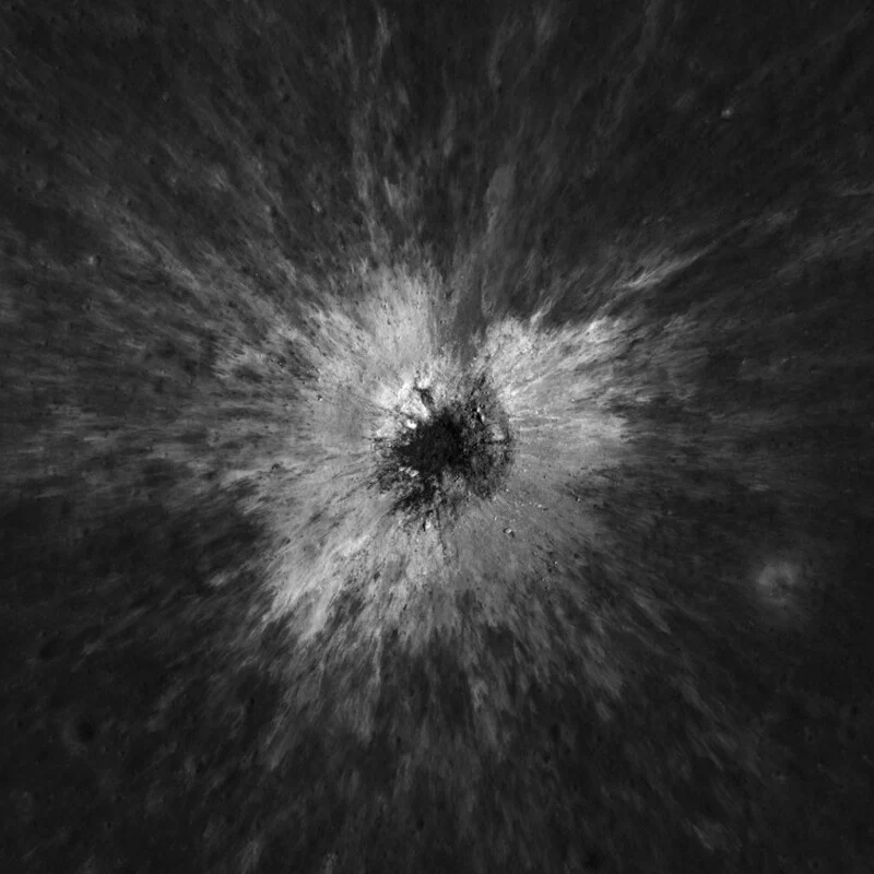 A high-contrast, overhead view of a lunar impact crater surrounded by a bright, radiating pattern of ejected material.
