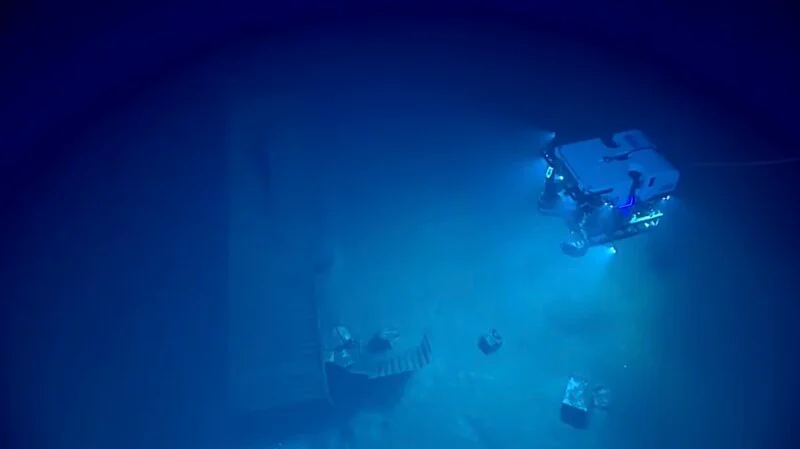 A remotely operated underwater vehicle illuminates the dark, deep-sea floor, where a wooden structure and scattered debris rest on the sediment.