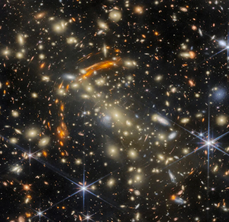 A dense galaxy cluster filled with numerous galaxies of varying shapes and sizes, featuring prominent gravitational lensing arcs.