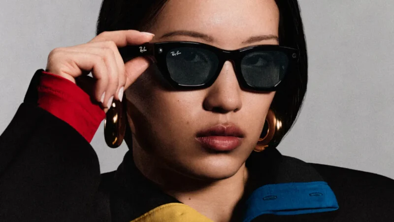 A person with dark hair wears black Ray-Ban sunglasses and large gold hoop earrings, with their hand resting on the frame.