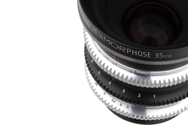 A close-up of a camera lens with silver and black gear-like rings, labeled "METAMORPHOSE 35 f/1.8," against a white background.