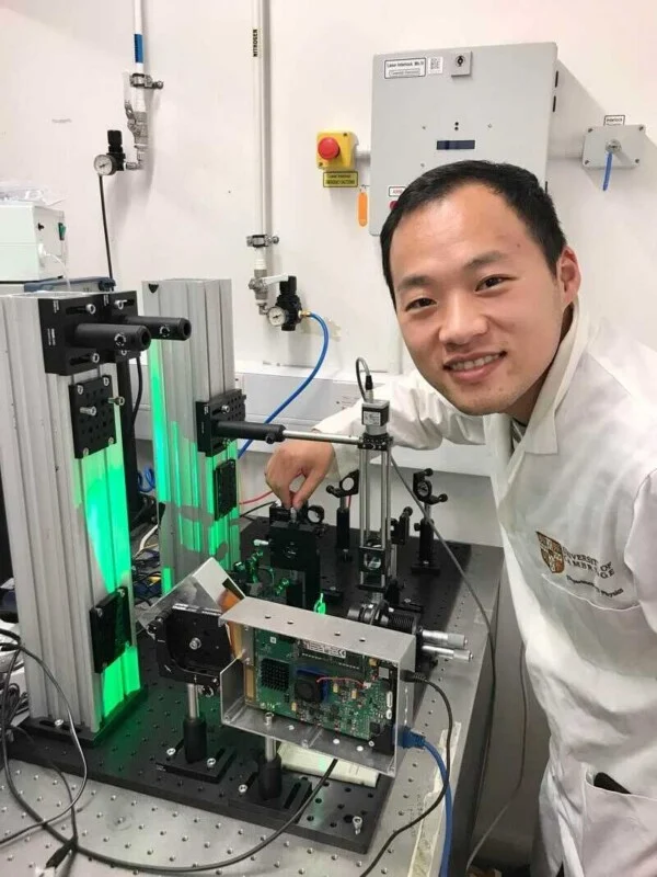 A smiling man in a white lab coat works with optical equipment and a circuit board on an experimental table in a laboratory.