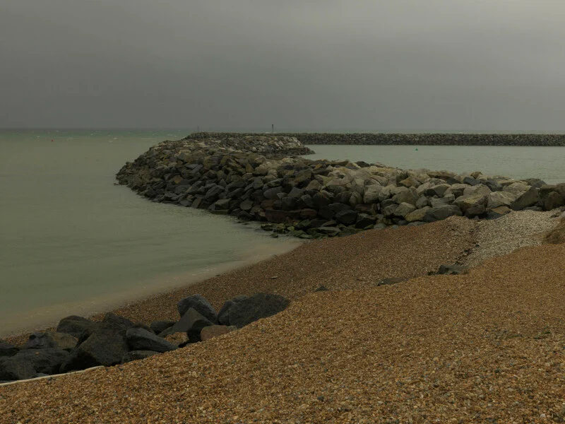 A curved stone breakwater extends into a calm, grey sea under an overcast sky, with a shingle beach in the foreground.