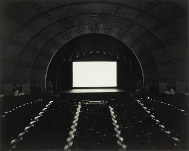 A wide-angle, black-and-white view of a large, arched theater interior with a brightly lit, blank movie screen.