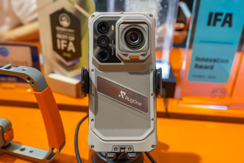 A Rugone rugged smartphone is mounted on a display stand, featuring a prominent camera module with multiple lenses and a textured light-gray back panel.