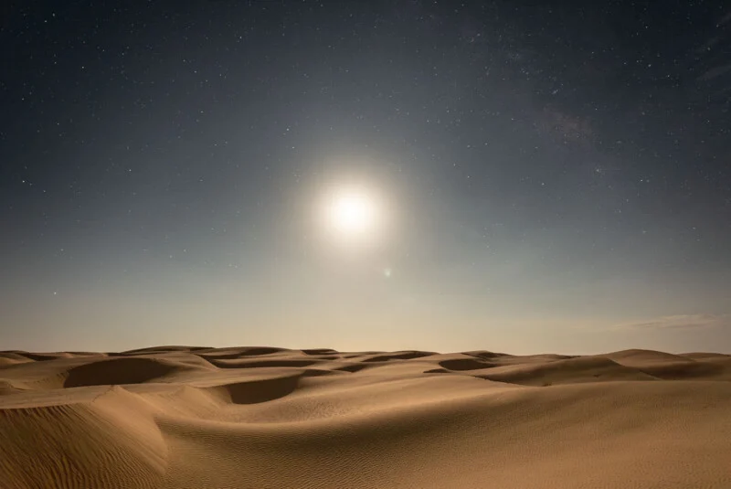 A bright moon illuminates a vast desert landscape with rolling sand dunes under a starry night sky.