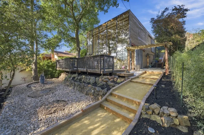 A modern two-story house with a glass-paneled facade stands on a hillside, featuring a wooden deck, a gravel garden, and a stepped dirt path leading to the entrance.