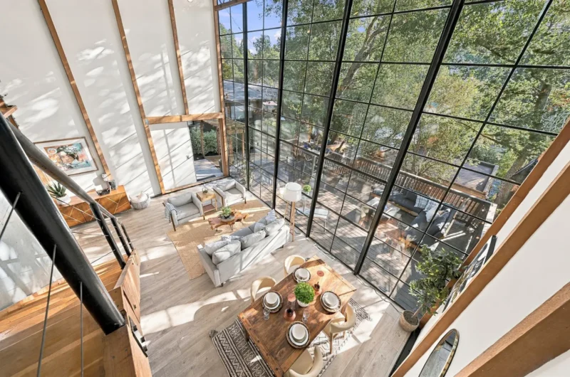 A high-angle view of a modern living and dining area with floor-to-ceiling windows, a wooden dining table, and a gray sofa set.