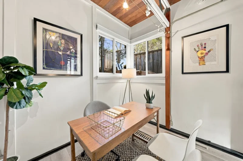 A home office features a wooden desk with a wire basket and an open book, flanked by two chairs, a floor lamp, and two framed artworks on white walls under a wood-paneled ceiling.