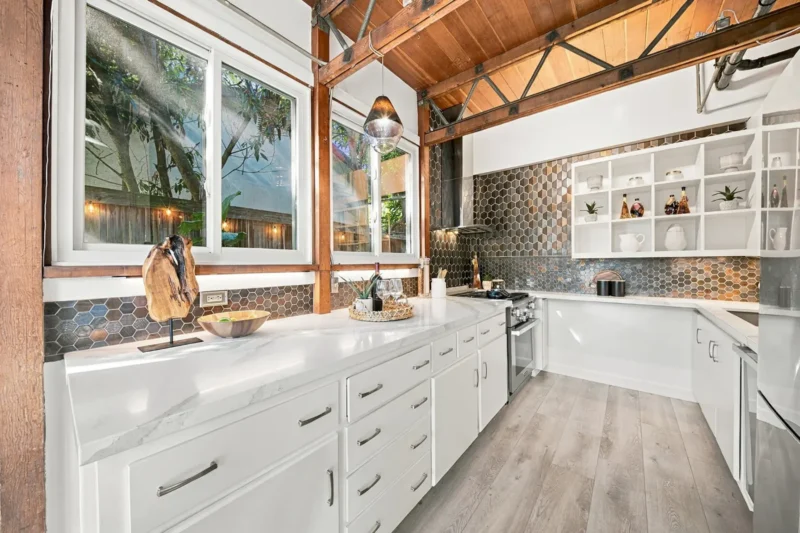 A modern kitchen features white cabinets, marble countertops, a hexagonal tile backsplash, and exposed wooden ceiling beams.
