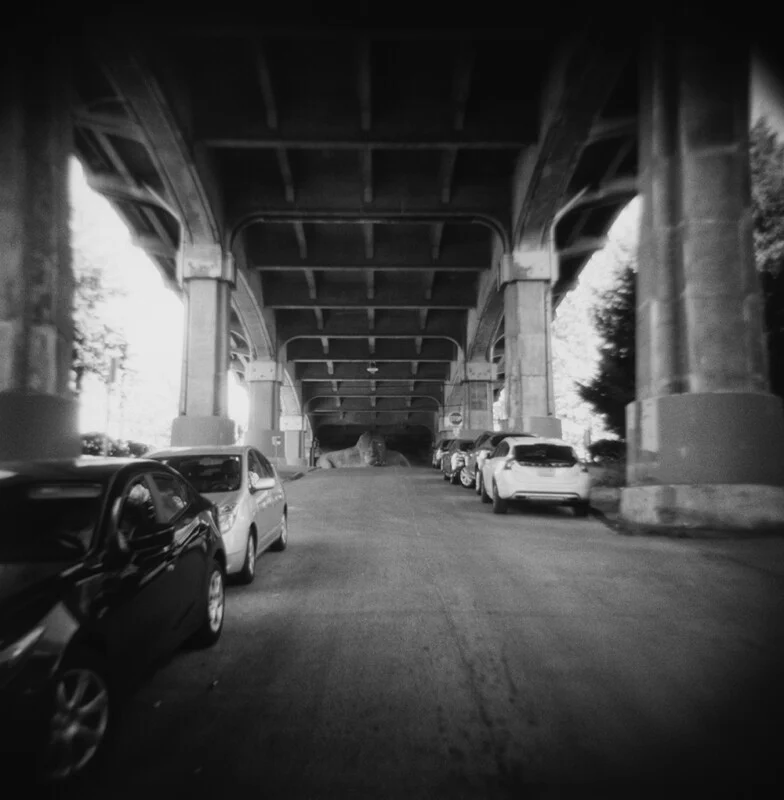 A black and white, wide-angle view looking down a road beneath a large concrete overpass, with parked cars lining both sides and a large, indistinct rock-like shape in the distance.