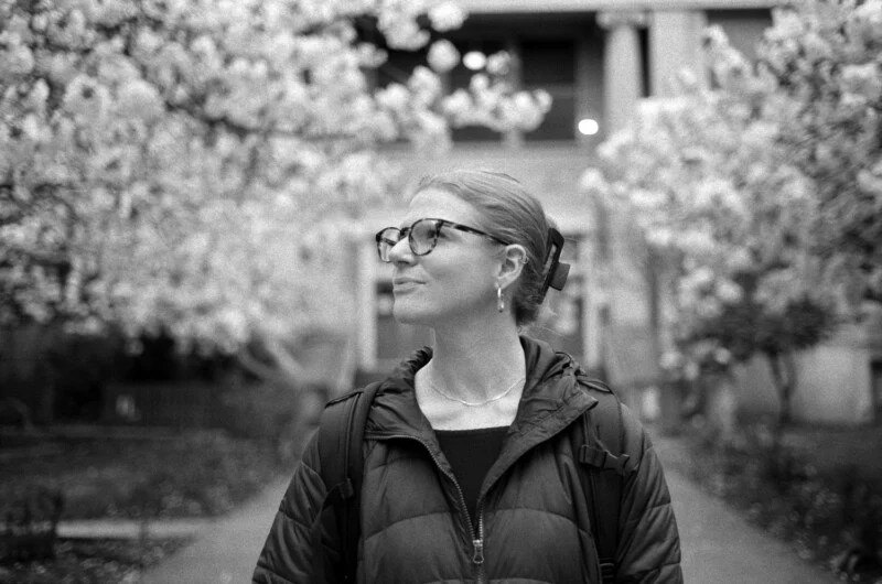 A person with glasses and hair pulled back looks to the side while standing outdoors between two flowering trees.