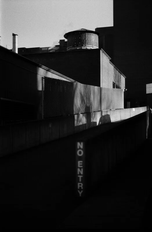 A black and white view of a concrete ramp with a vertical "NO ENTRY" sign, leading toward a brick building with a water tower.