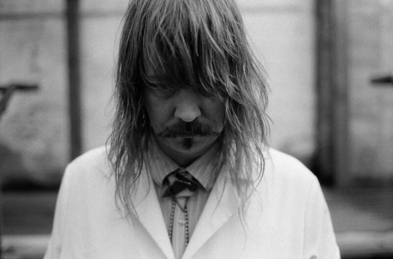 A black-and-white portrait of a person with long hair and a mustache, looking downward while wearing a collared shirt and a white jacket.
