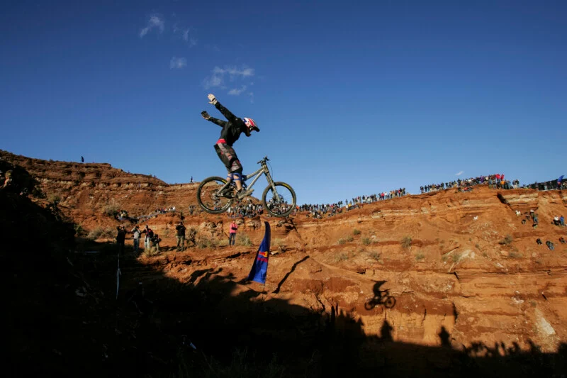 A mountain biker performs a no-handed jump in mid-air above a rocky canyon with spectators watching from the ridge.