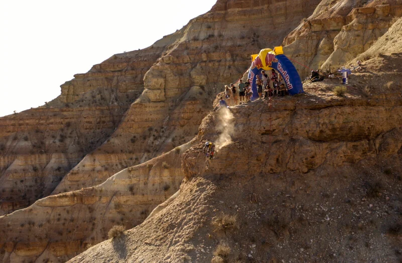 A mountain biker descends a steep, rocky cliff face below a Red Bull branded inflatable arch where a group of spectators is gathered.