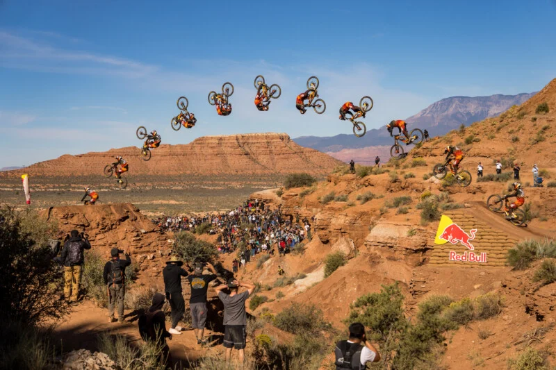 A composite action shot shows a mountain biker performing a backflip over a desert canyon, with a crowd of spectators watching from below.