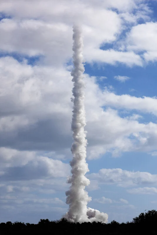 A thick, vertical column of white smoke rises from behind a dark treeline into a blue sky filled with scattered clouds.