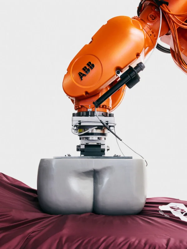 An orange ABB industrial robotic arm holds a gray, buttocks-shaped object over a dark red fabric surface.