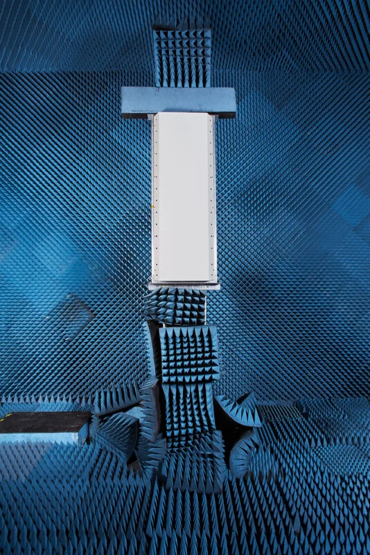 A rectangular white device is mounted vertically in the center of an anechoic chamber lined with blue, pyramid-shaped foam absorbers.