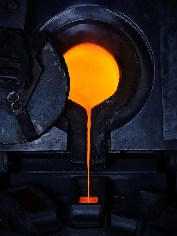 Molten metal glows bright orange as it pours from a dark industrial furnace into a rectangular mold below.