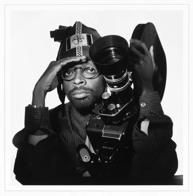 Spike Lee poses in a black-and-white portrait, wearing a branded hat and glasses while holding a Bolex movie camera.