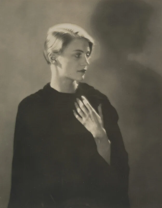 A black-and-white portrait of a person with short blonde hair, wearing a dark garment and holding one hand to their chest, set against a soft, shadowed background.