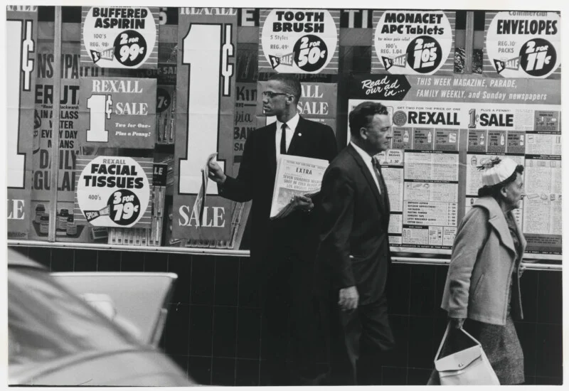 Malcolm X walks past a Rexall store window filled with "1¢ Sale" advertisements, holding a newspaper with a headline about a police shooting.